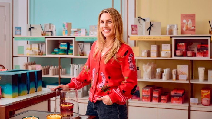 Nicola Elliott stands smiling in the NEOM wellbeing store, surrounded by NEOM products on display shelves.