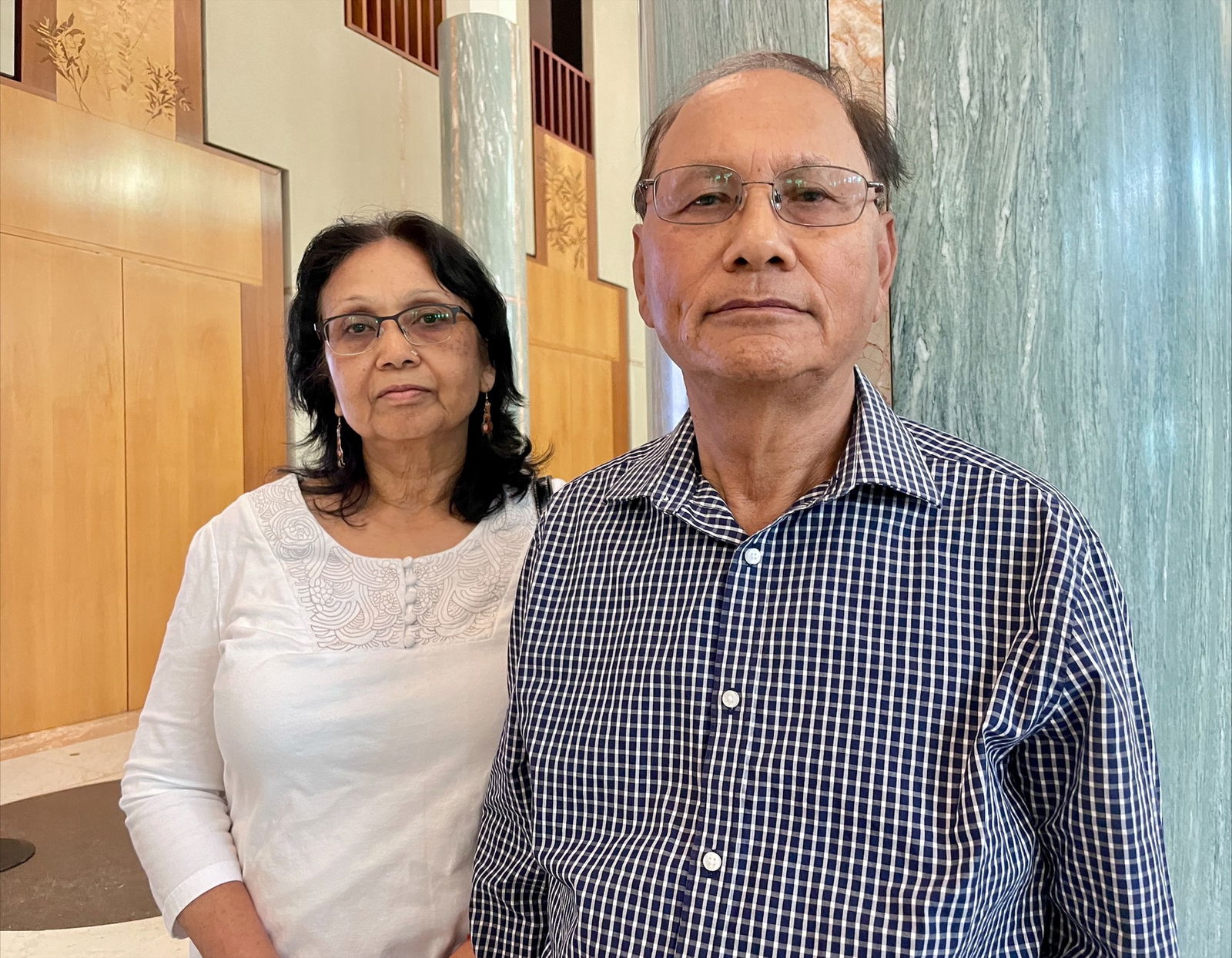 A dark haired woman in a white shirt stands with a dark haired man in a blue and white checked shirt.