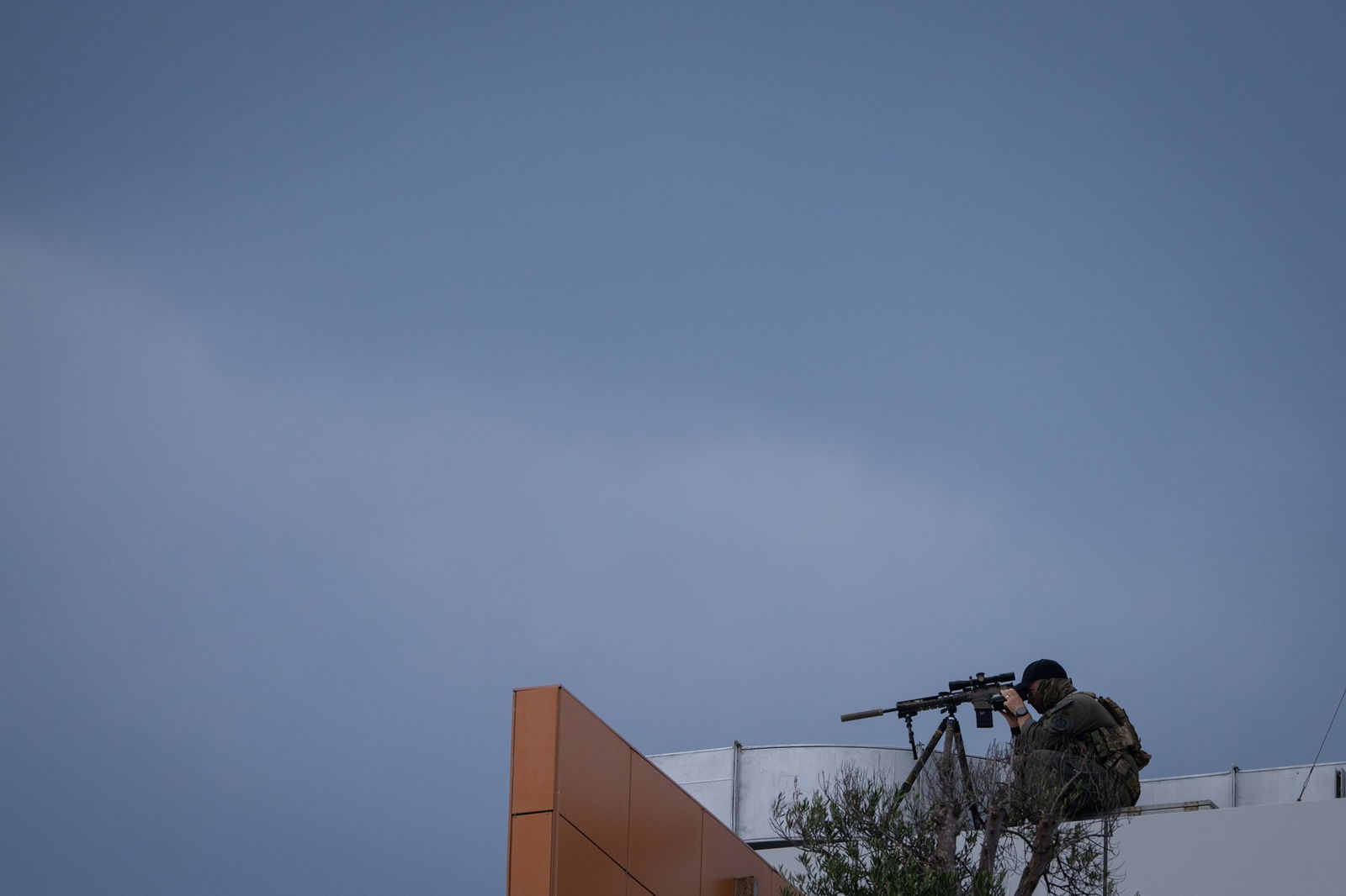 A man in dark clothing is poised with a sniper on the top of a building.