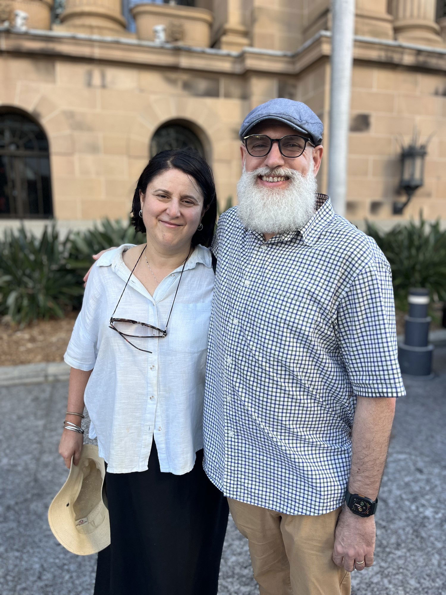 A dark haired woman in a white shirt and a white bearded man in a blue cap, glasses and blue checked shirt stand outside a sandstone building.