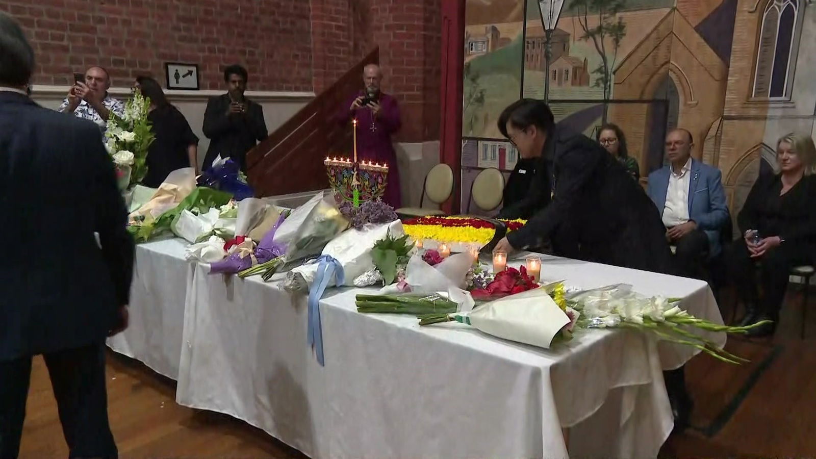 Bouquets of flowers lie on a table as people gather round.