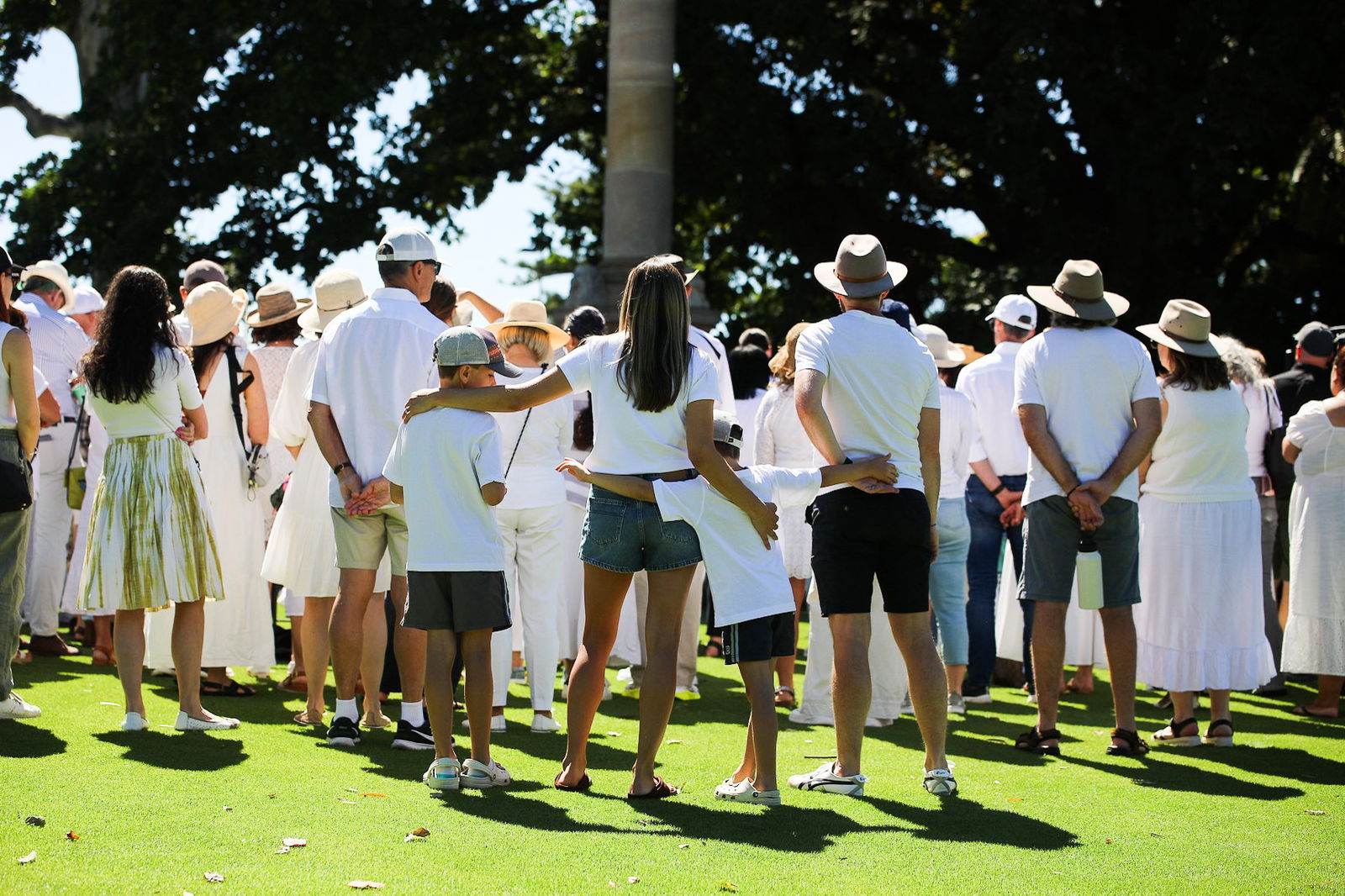 A large group of people stand in a park facing away from the camera with their arms around each other. 