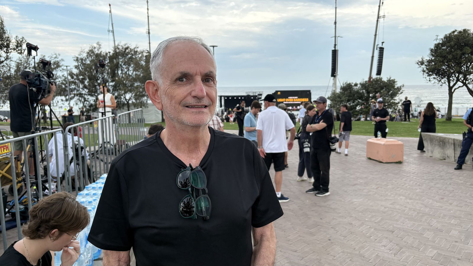 Man in black shirt smiles with media set up behind him at Bondi Beach