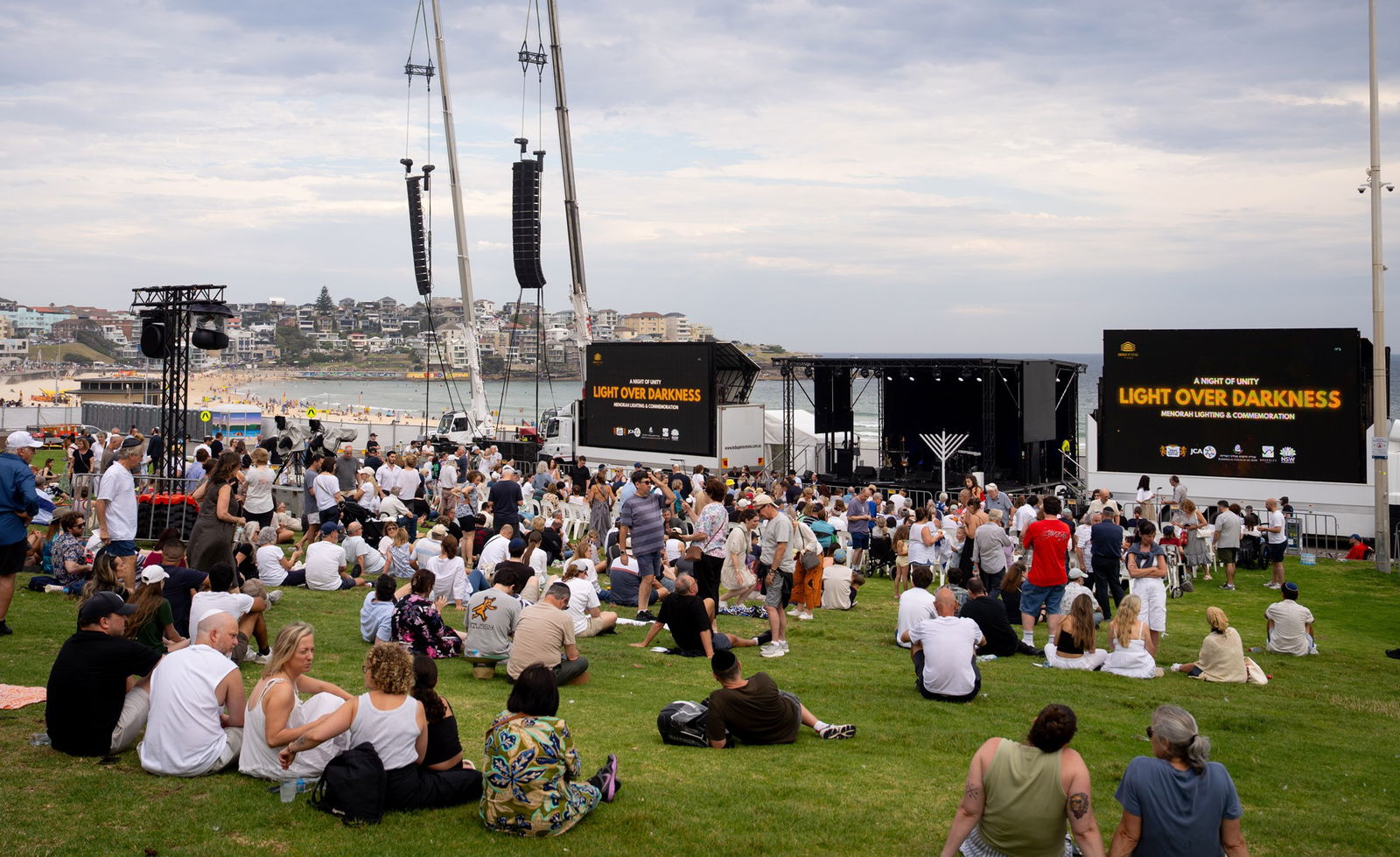 A crowd of people sits on a grassy hill overlooking Bondi Beach looking at large screens.