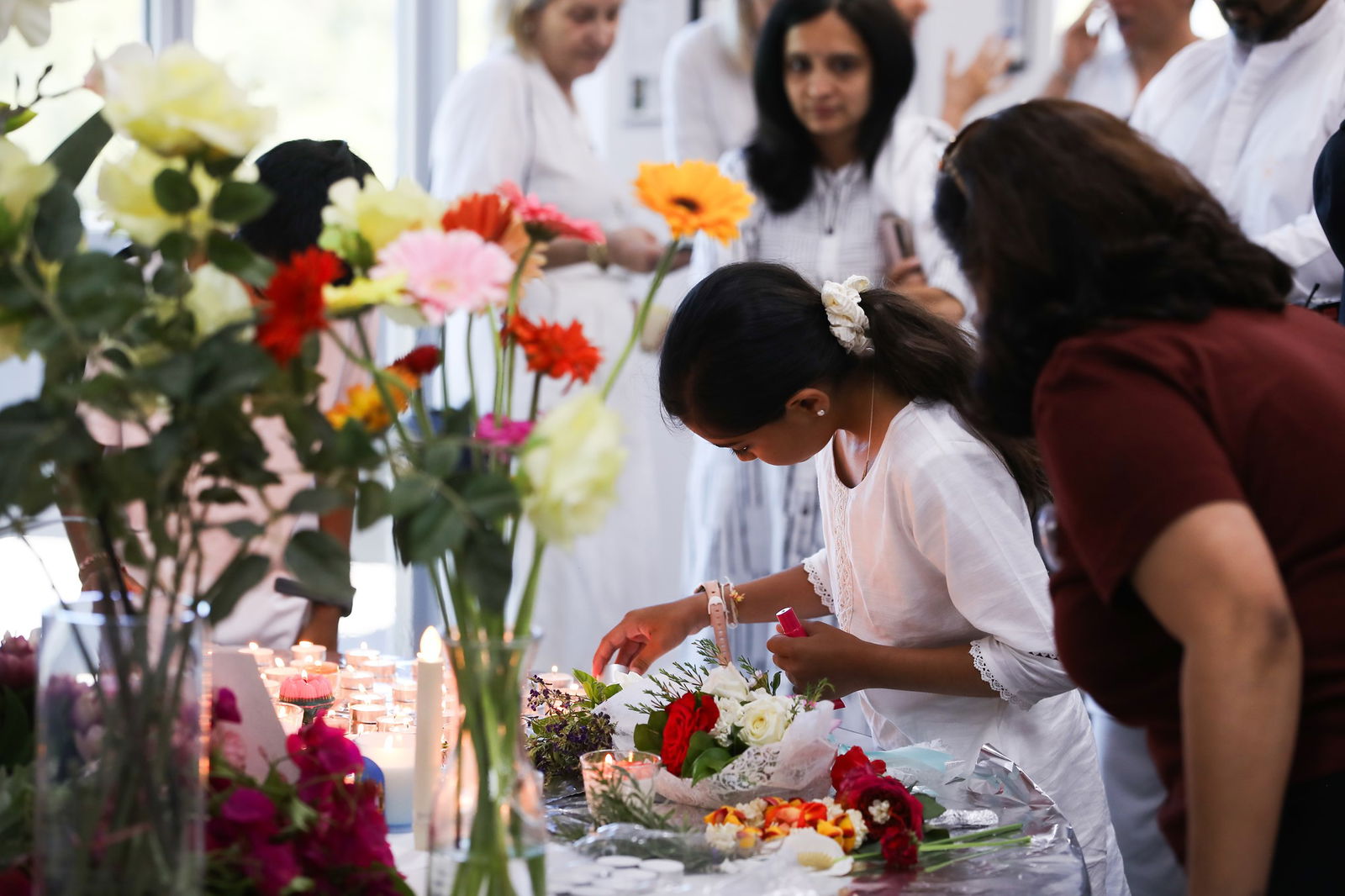 A young girl lights a candle on a table covered in flowers. She is surrounded by other women dressed in white. 