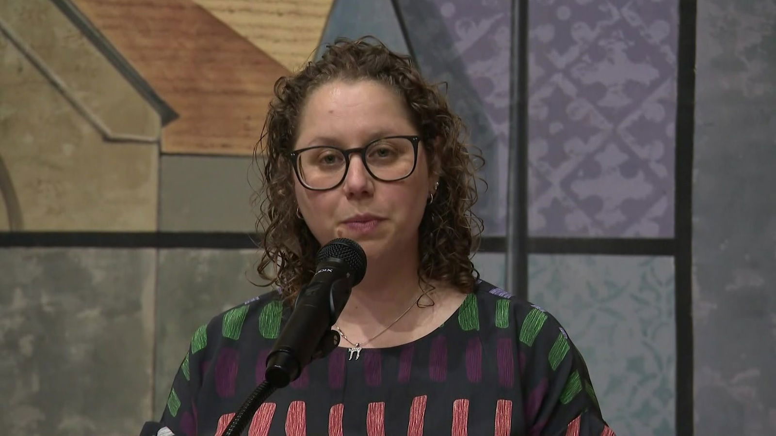 A woman with curly hair and glasses in a black top with colourful rectangles speaks into a microphone.
