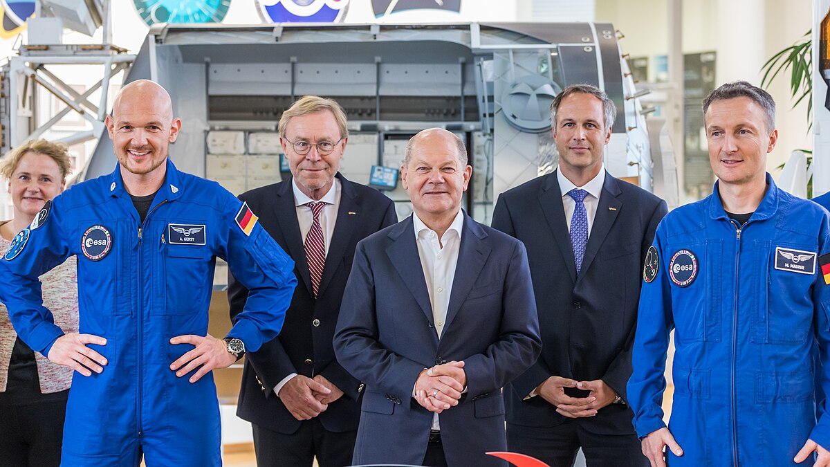 Two men in blue flight suits stand on either side of a man in a suit and tie with men in suits behind him