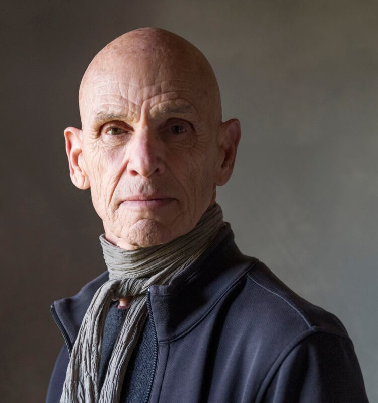 An older bald man with a serious expression wears a textured gray scarf and a dark zip-up jacket, standing against a plain, softly lit background.