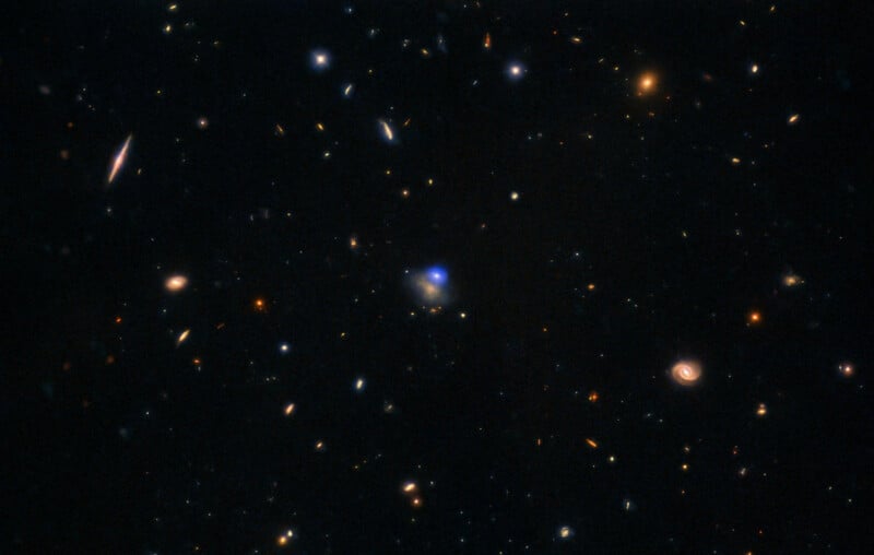 A deep space image showing numerous distant galaxies of various shapes and colors scattered across a dark background, with a bright blue central spot.