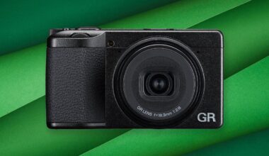 A compact black digital camera with a prominent GR lens is centered on a background of overlapping green paper strips.