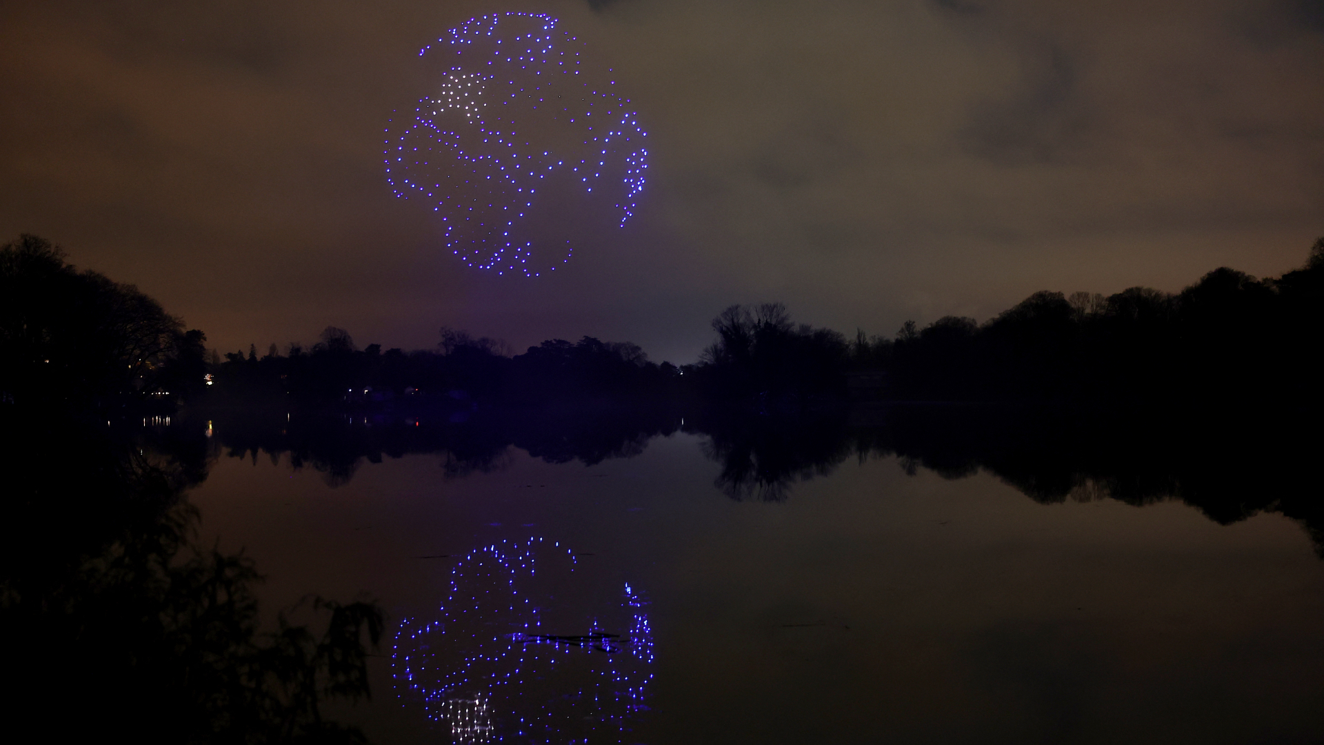 A series of purple drones come together in the night sky above a still lake to create a 3D image of planet Earth with the continent of Africa seen and reflected in the lake