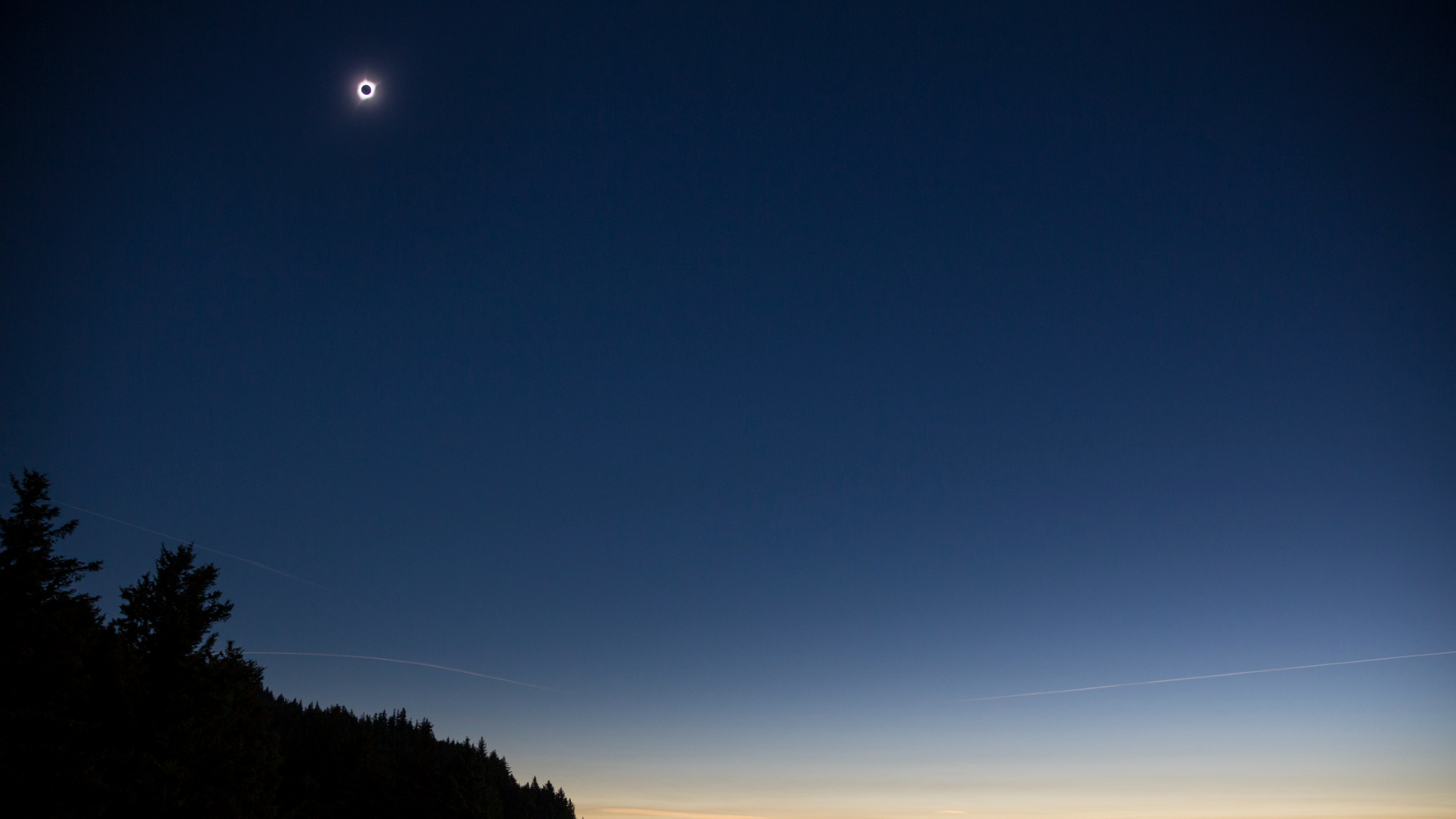 A large glowing halo for a solar eclipse is seen in a dark blue night sky with a silhouetted landscape in the front of the image