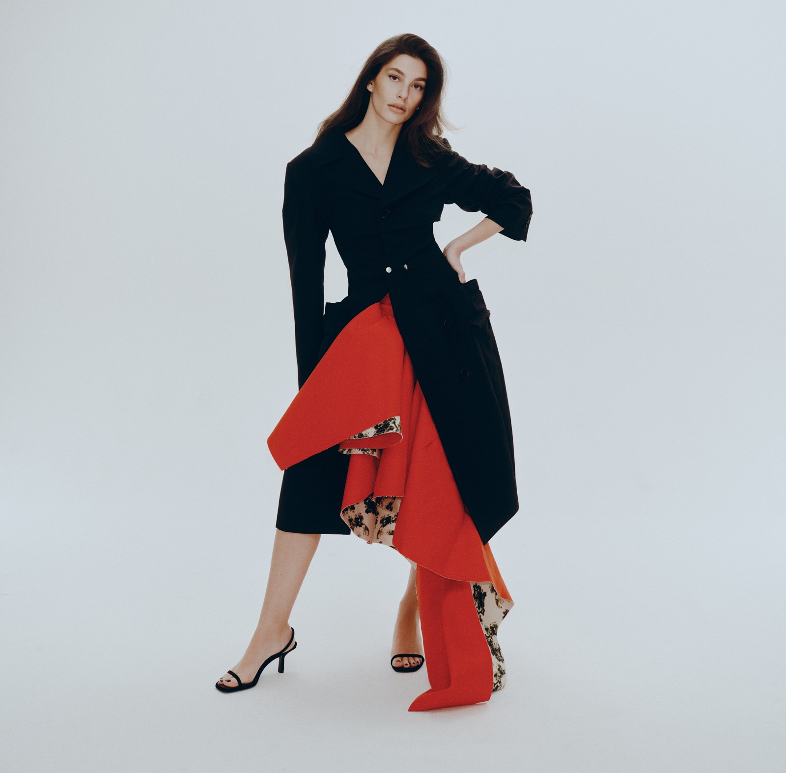 Camila Morrone wearing a black coat over a red and patterned skirt.