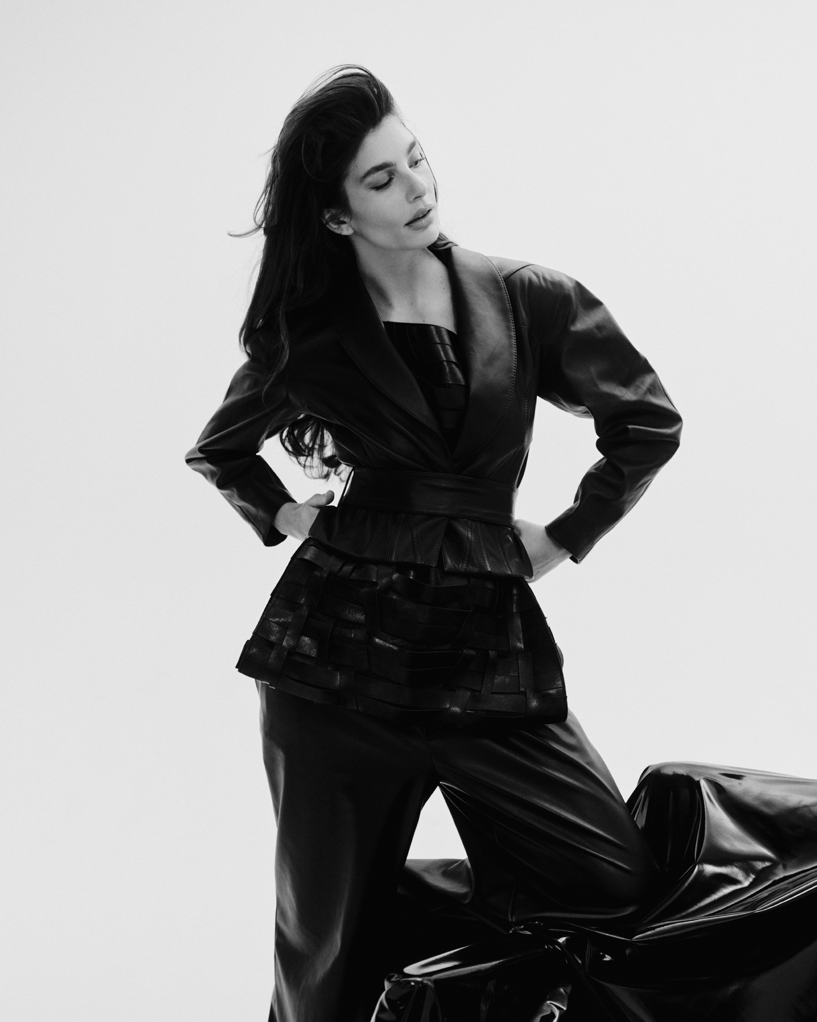 Camila Morrone in a black leather outfit, looking to the side with her hands on her hips.