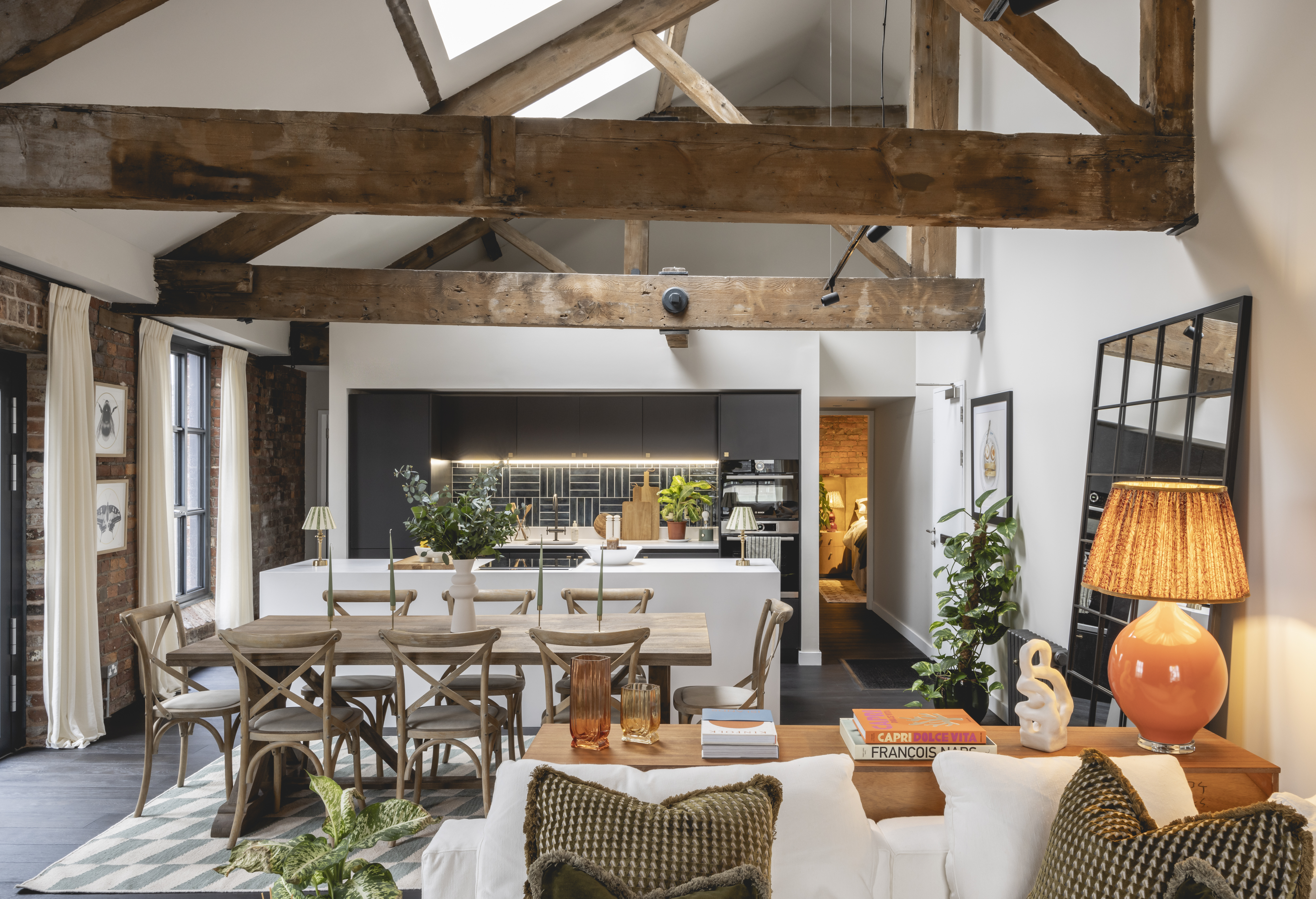 A living room with a dining area and kitchen under a high ceiling with exposed wooden beams, white walls, and skylights.