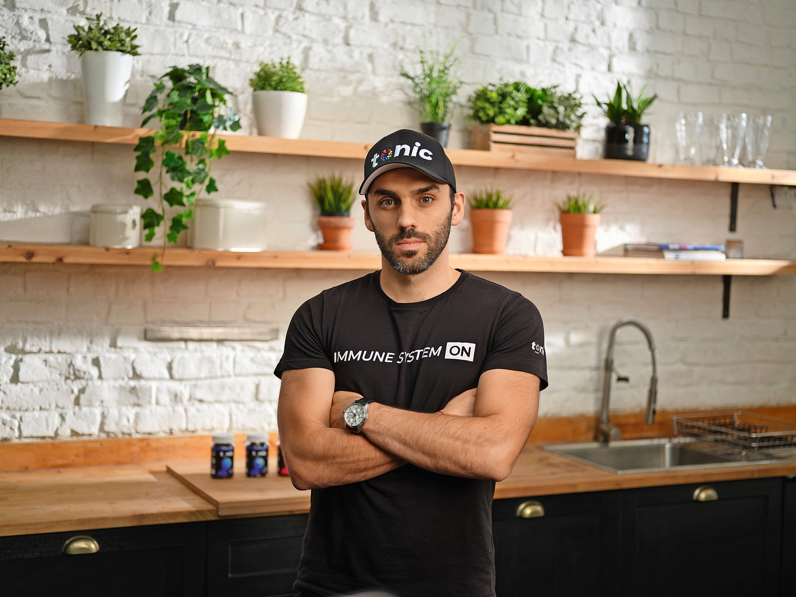 Man in a "Tonic" cap and "IMMUNE SYSTEM ON" shirt stands in a kitchen with bottles on the counter.