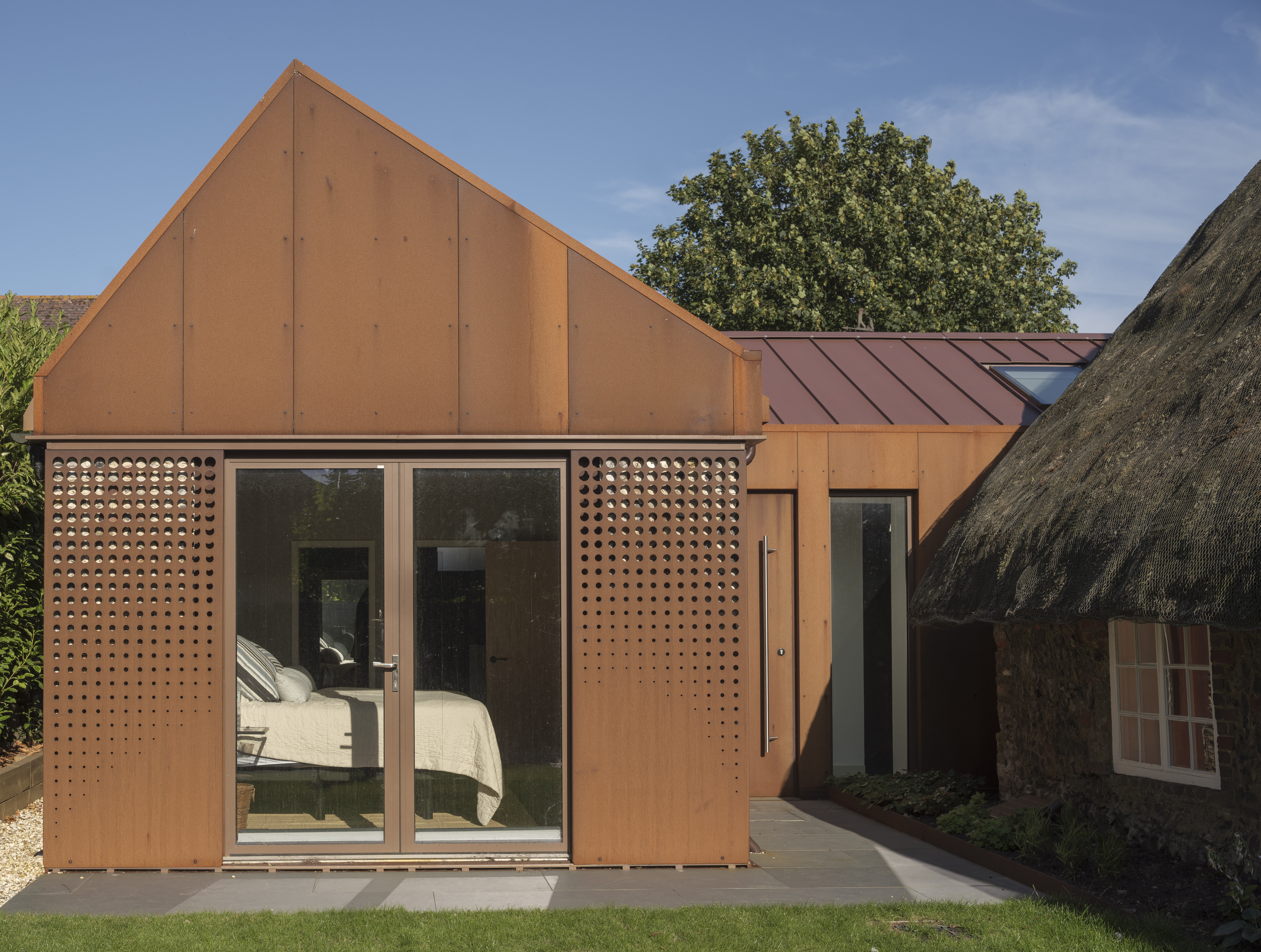 A modern cottage with rusty orange walls and a gabled roof, alongside an older building with a thatched roof, in Laurel Cottage.