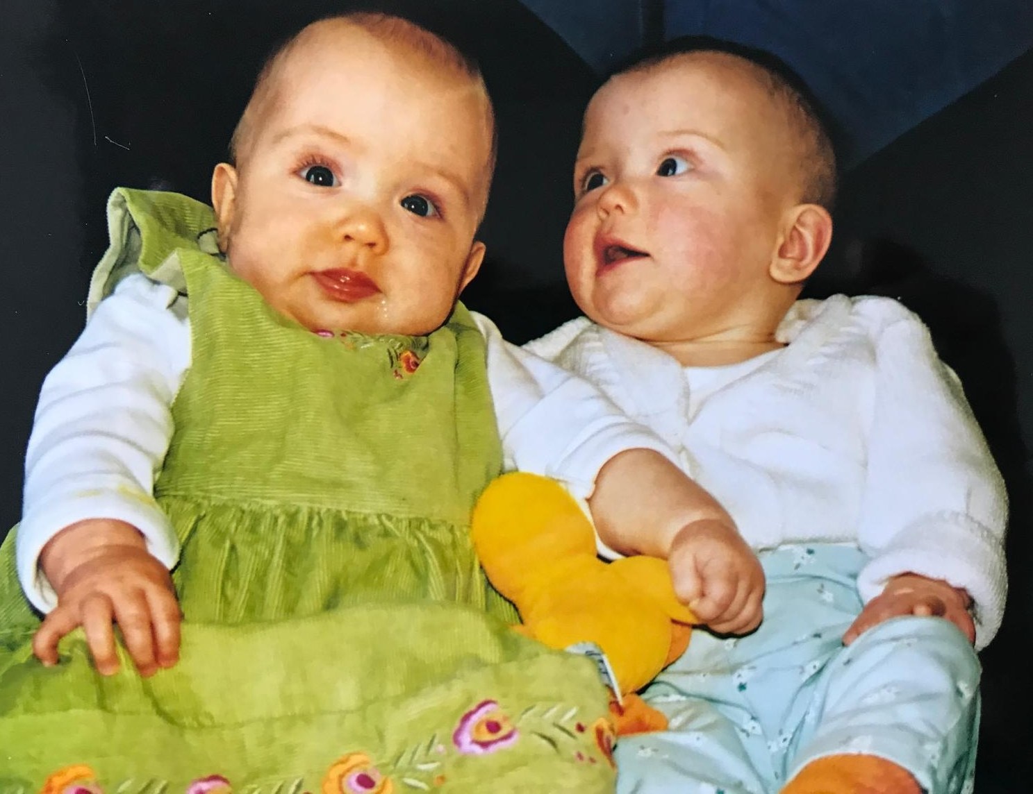 Two babies sitting together, one in a green dress and the other in a white top and blue pants holding a yellow toy.