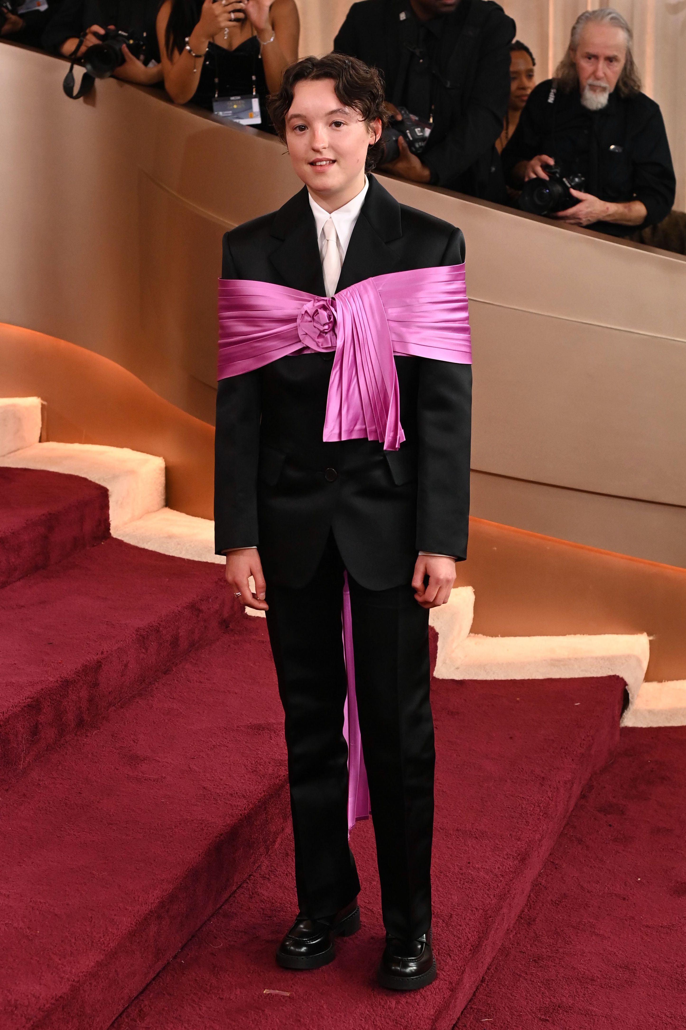 Bella Ramsey in a black suit with a large pink sash wrapped around their torso and a floral embellishment.