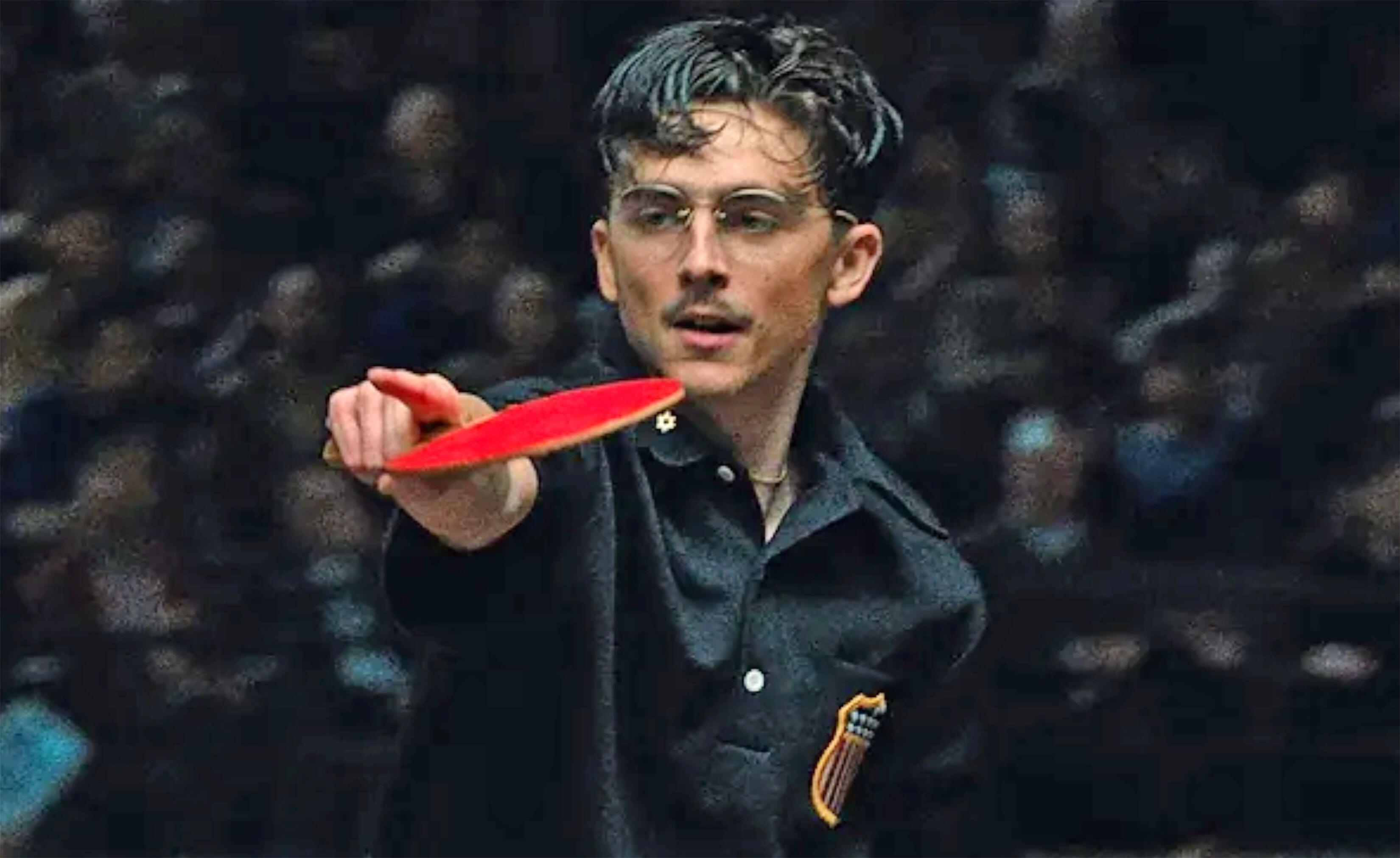 Timothée Chalamet, as Marty Supreme, holds a red ping pong paddle.