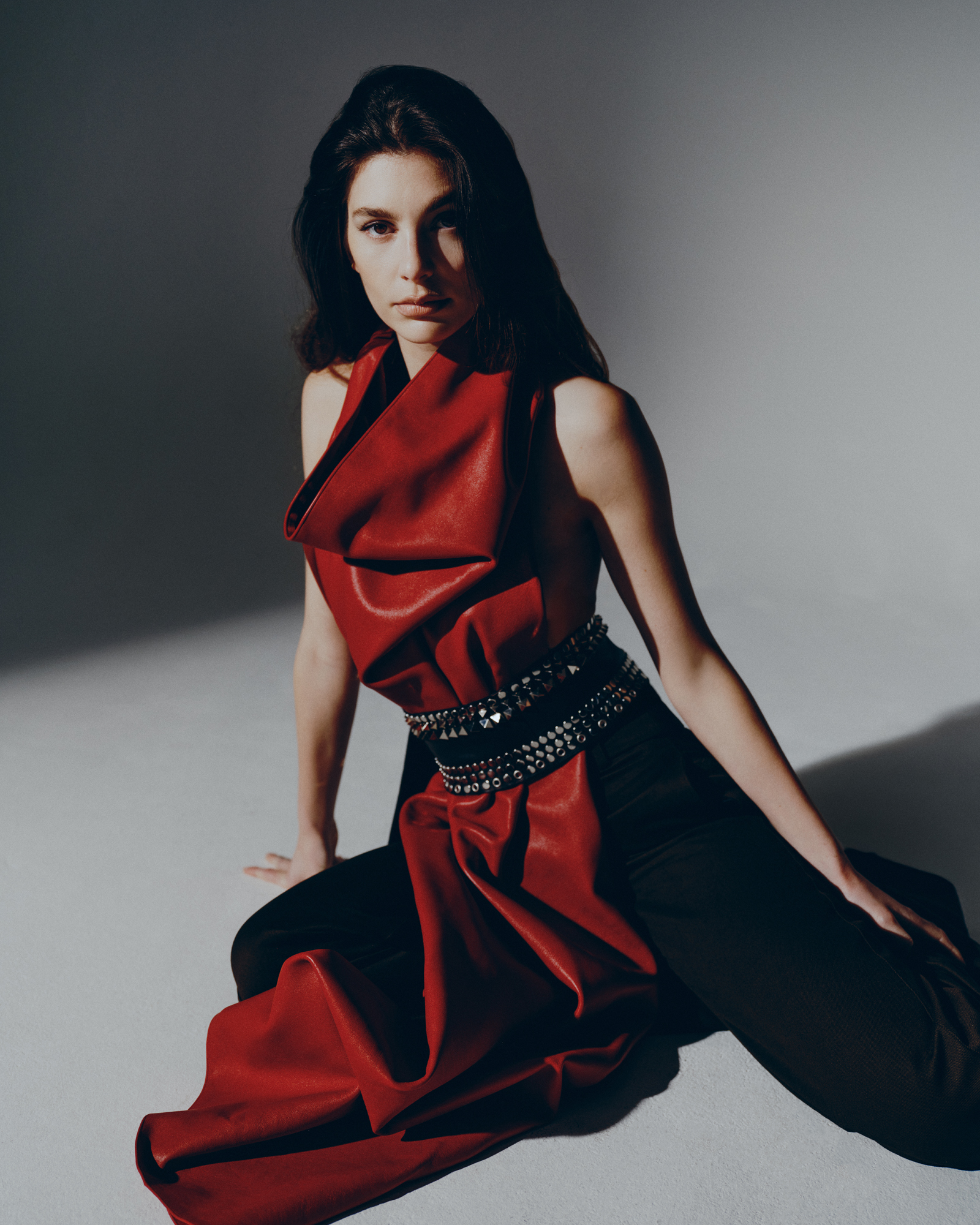 Camila Morrone sitting on the floor in a red top, black pants, and a studded belt.