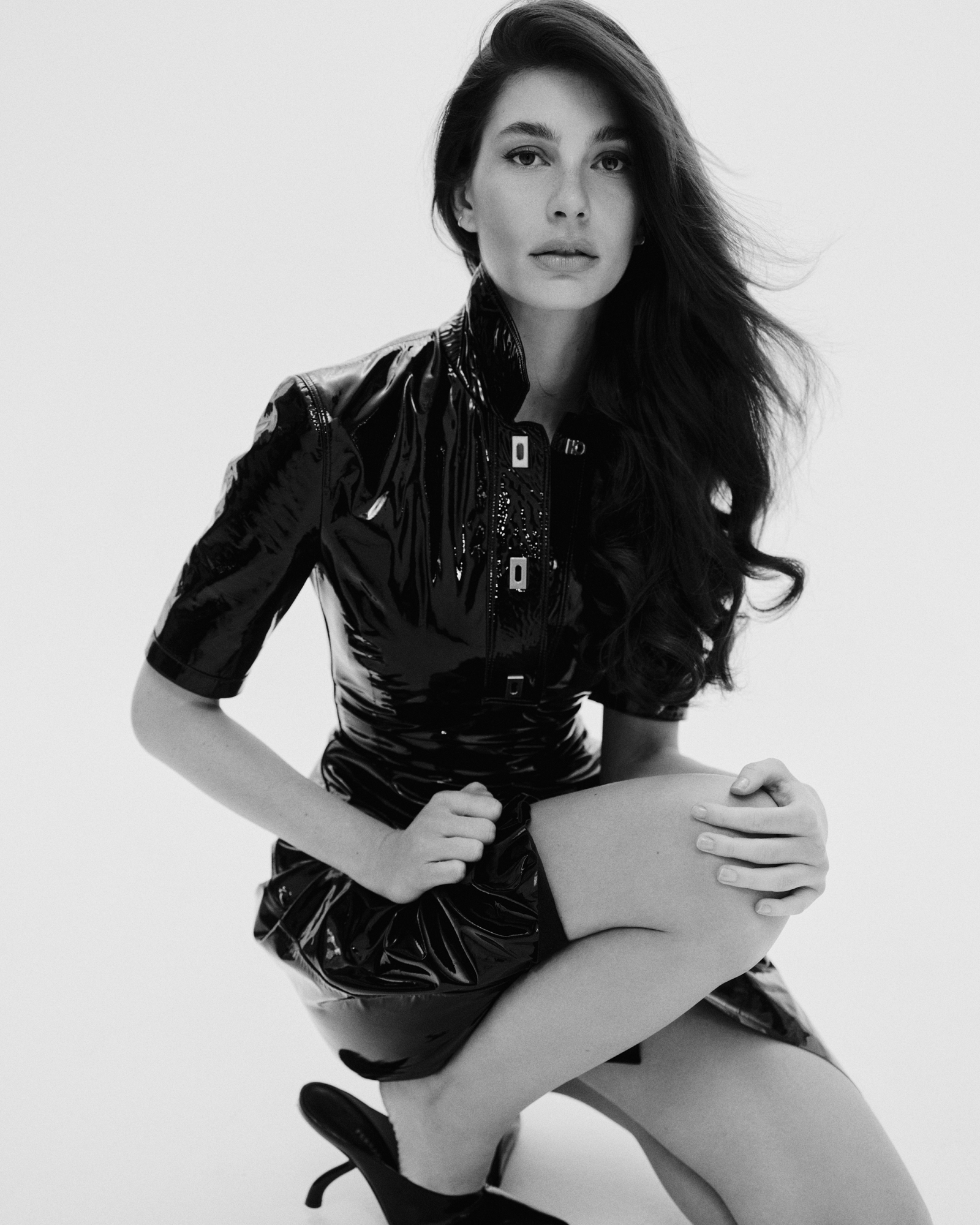 Camila Morrone in a shiny black dress with a high collar, kneeling.