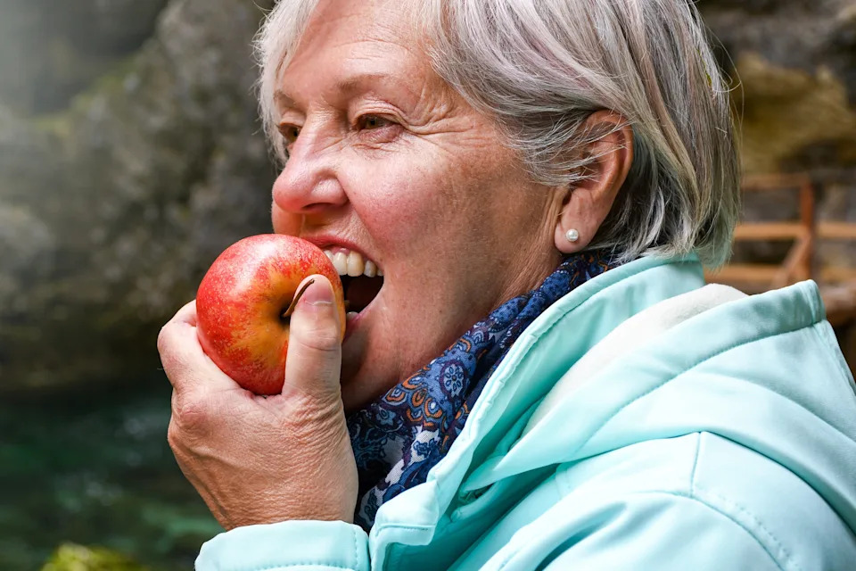 A person enjoys biting into an apple outdoors, wearing a scarf and jacket, suggesting a brisk environment