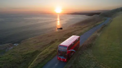 Getty Images Red bus being driven along a quiet coastal road with the sun setting behind it.