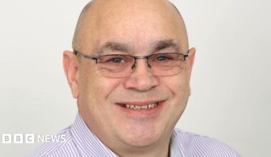 A woman wearing a blue hoodie and a purple jacket with her face buried in her hands, standing in an alleyway.