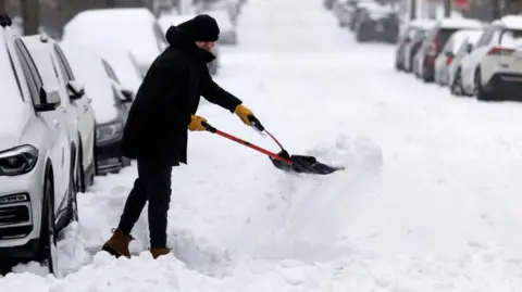 Will Oliver/EPA/Shutterstock A man rugged up in heavy winter clothing uses a tool to clear snow packed into a shovel he is using to clear part of a road near a line of parked cars, all of which have snow over their roofs and windscreens.