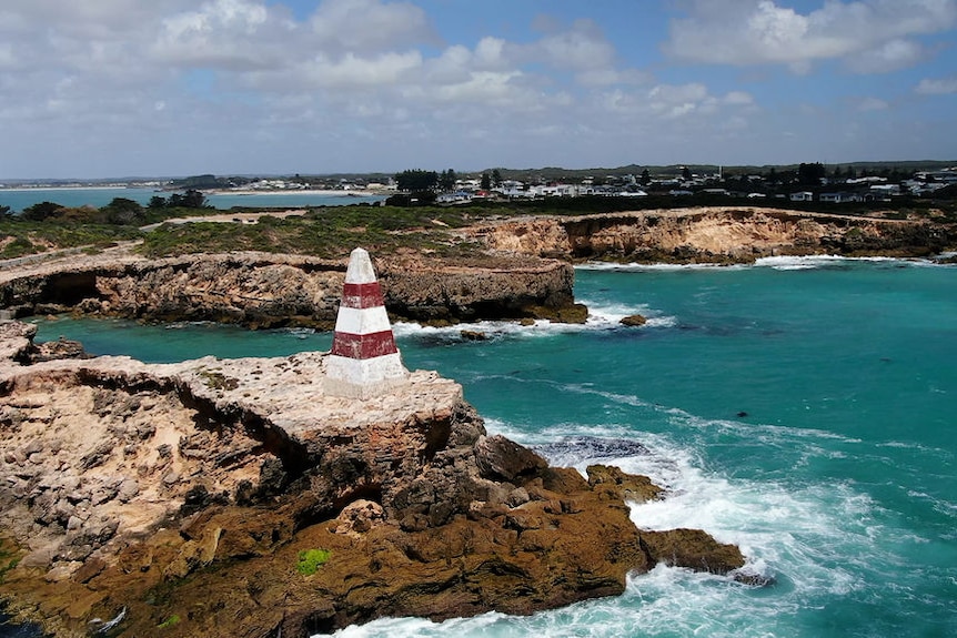 A red and white obelisk sitting on a cliff, with waves crashing into the side of the cliff.
