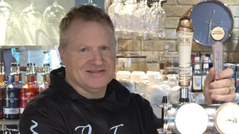 James Graham James stands behind the bar pulling a pint while facing the camera. In the background are gin bottles in different colours and stacked white tea cups. He is wearing a black hoodie with white writing on it saying the name of his pub, and has short dark hair.