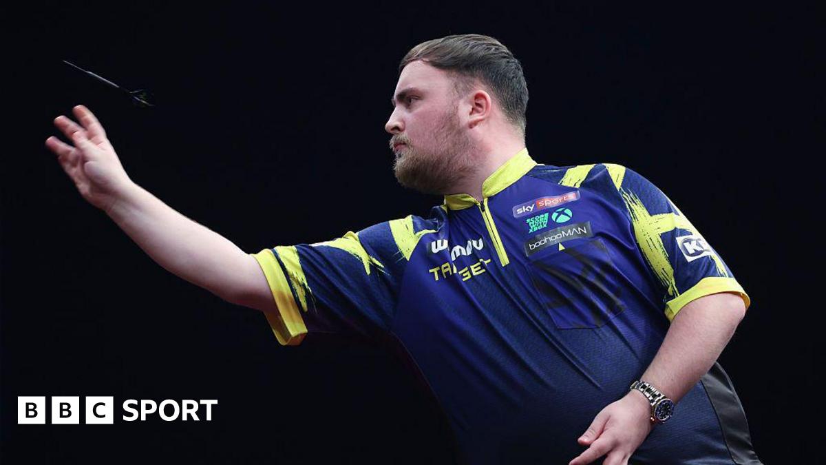 A side-on view of Luke Little as he throws a dart. He has dark hair, brushed  forward, and a beard. His shirt is blue with yellow trim.