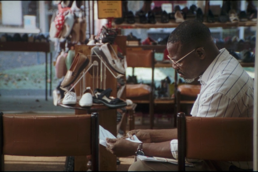 A Black man sitting in a shoe shop looking at a notice.