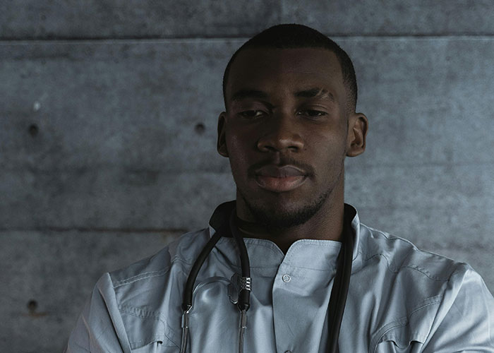 Young male medical professional in white coat with stethoscope, reflecting on worst things said to patients in healthcare settings