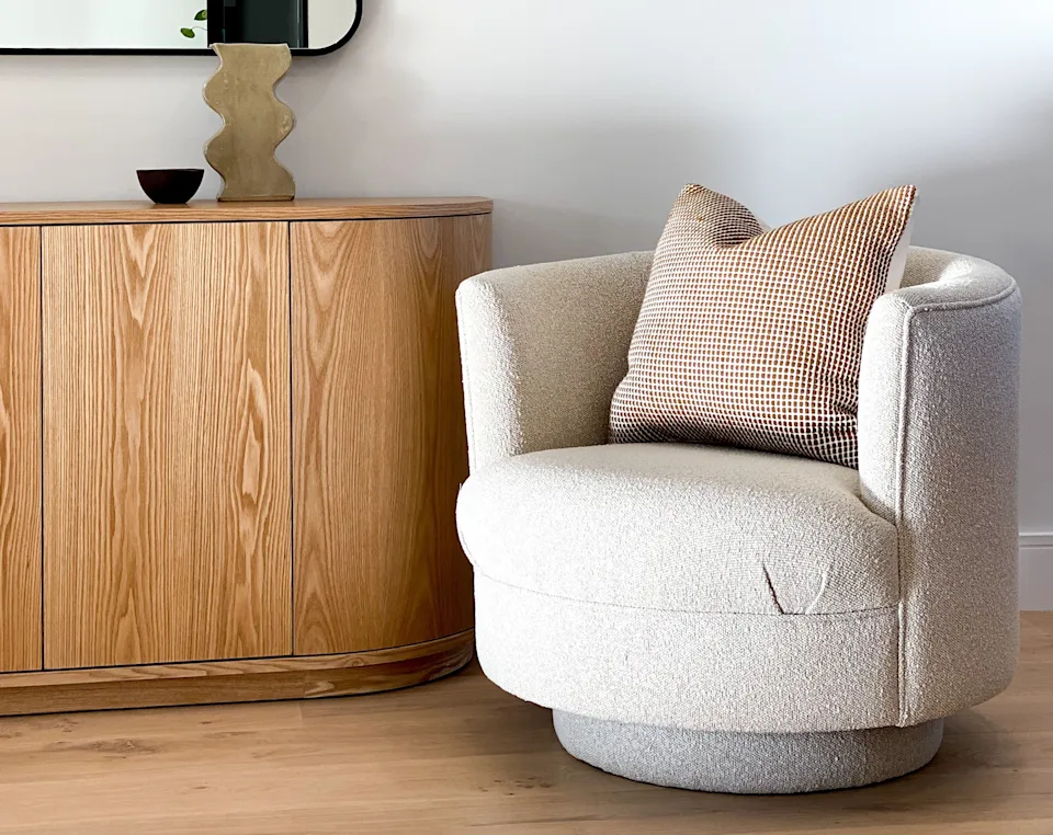 Stylish modern living room with a curved wooden sideboard, decorative vase, round mirror, and cozy armchair with a checkered cushion