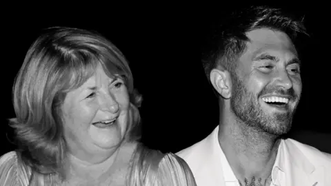 The Couture Club Ross Worswick and his mother Judith are sitting side by side and are laughing while staring off camera in this black and white photograph.