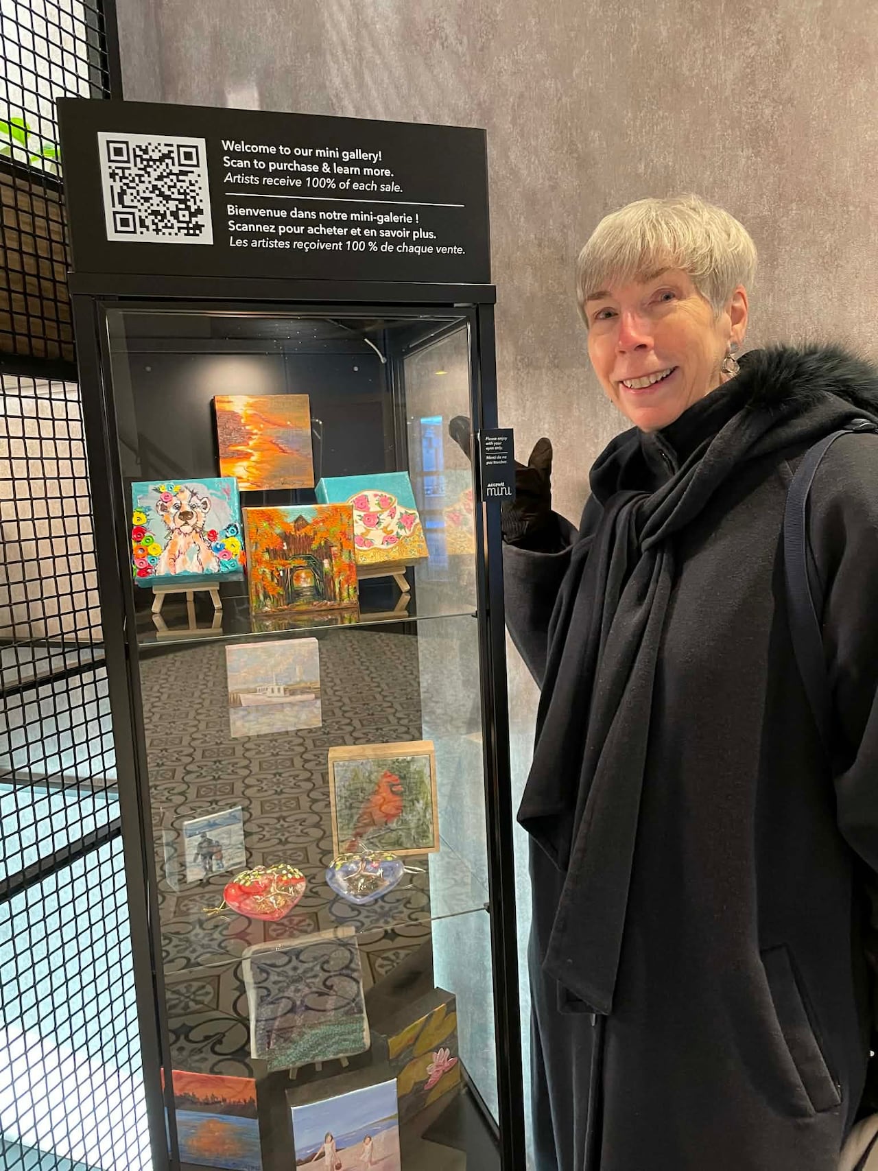 A white woman with white hair, she is pointing toward a painting in a rectangular glass shelf filled with mini artwork.
