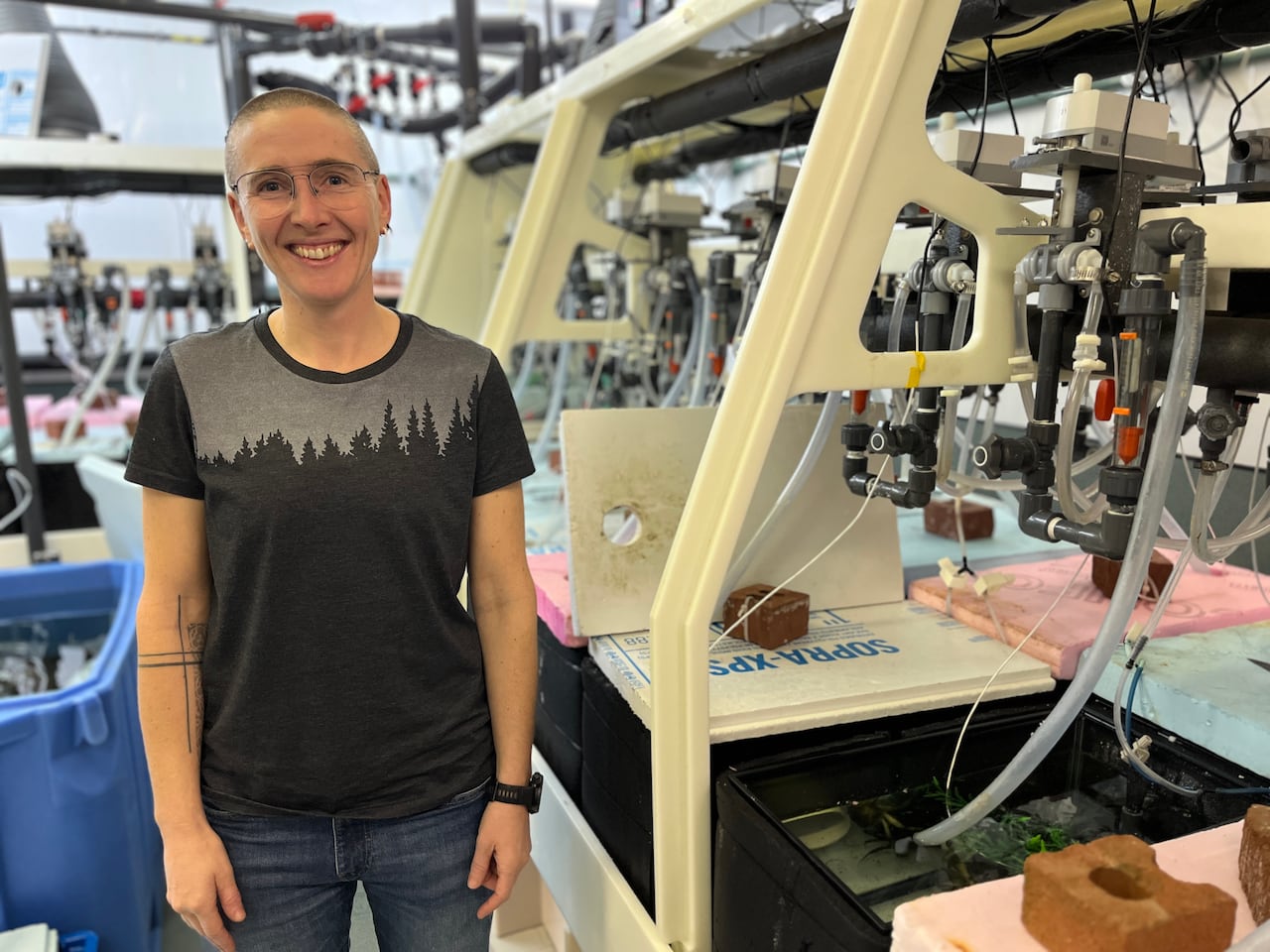 A woman in a grey and black t-shirt, with a tattoo along her arm, stands in a lab. 