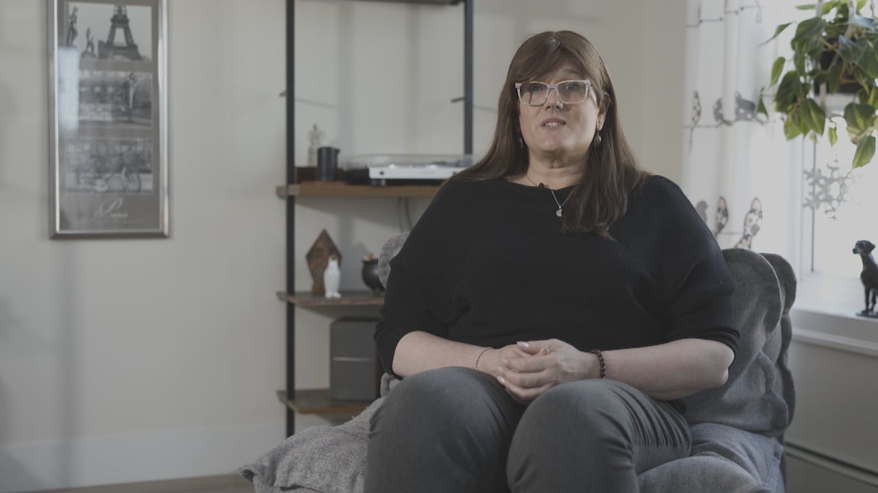 A woman in a black top sits in a chair and speaks to the camera.