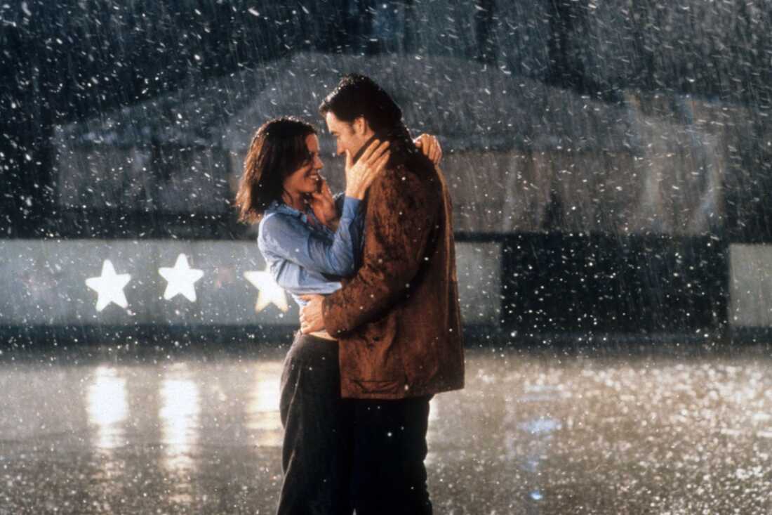 Actors Kate Beckinsale and John Cusack embrace one another in the rain in the 2001 film Serendipity. Her hands are around his shoulders, and his hands are around her waist.