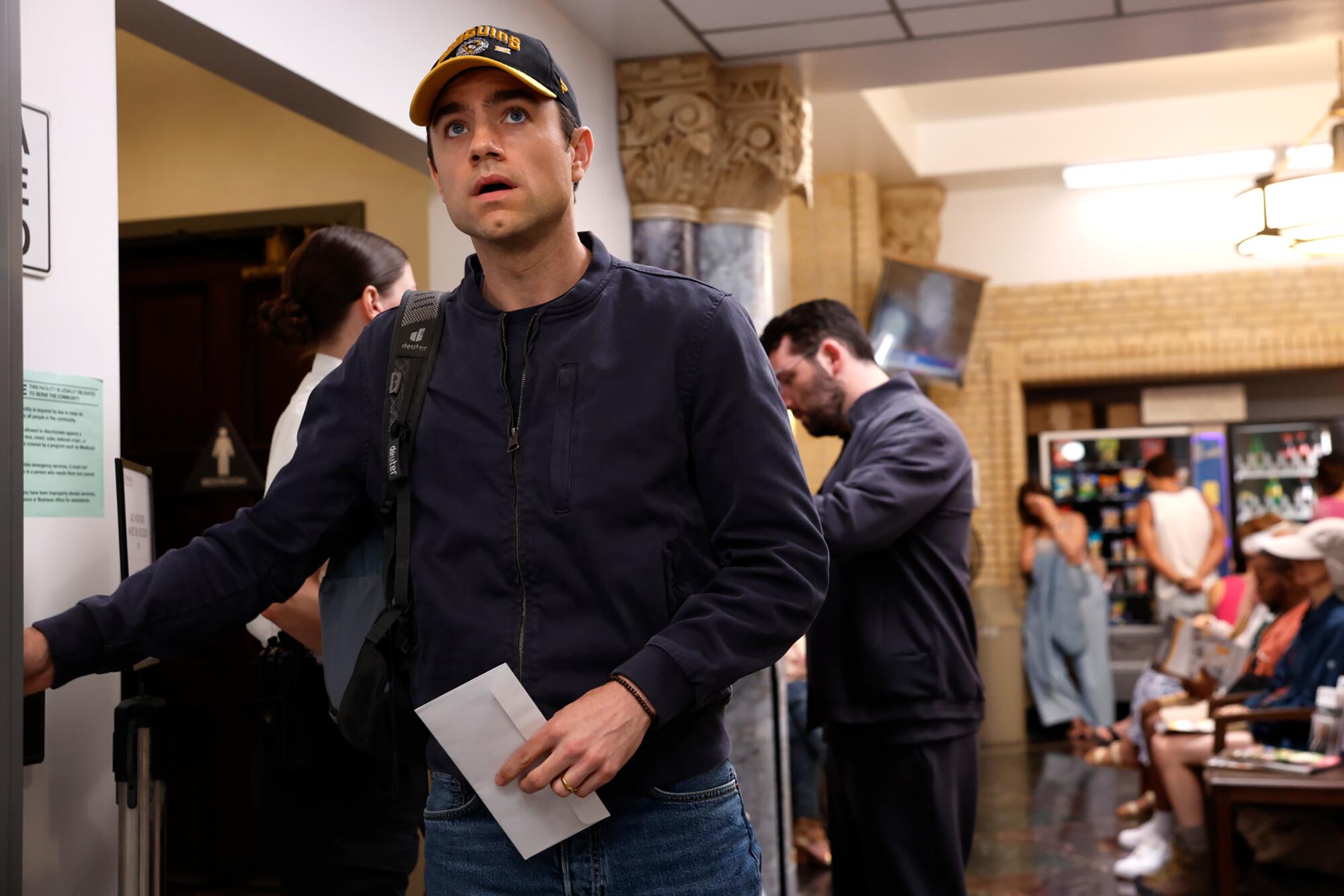 A man wearing a baseball cap walks with an envelope in his left hand