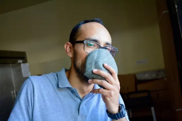 A man with medium skin tone, wearing glasses and a light gray polo shirt, holds up a blue tortilla up to his face, partially covering it from his nose to his mouth.
