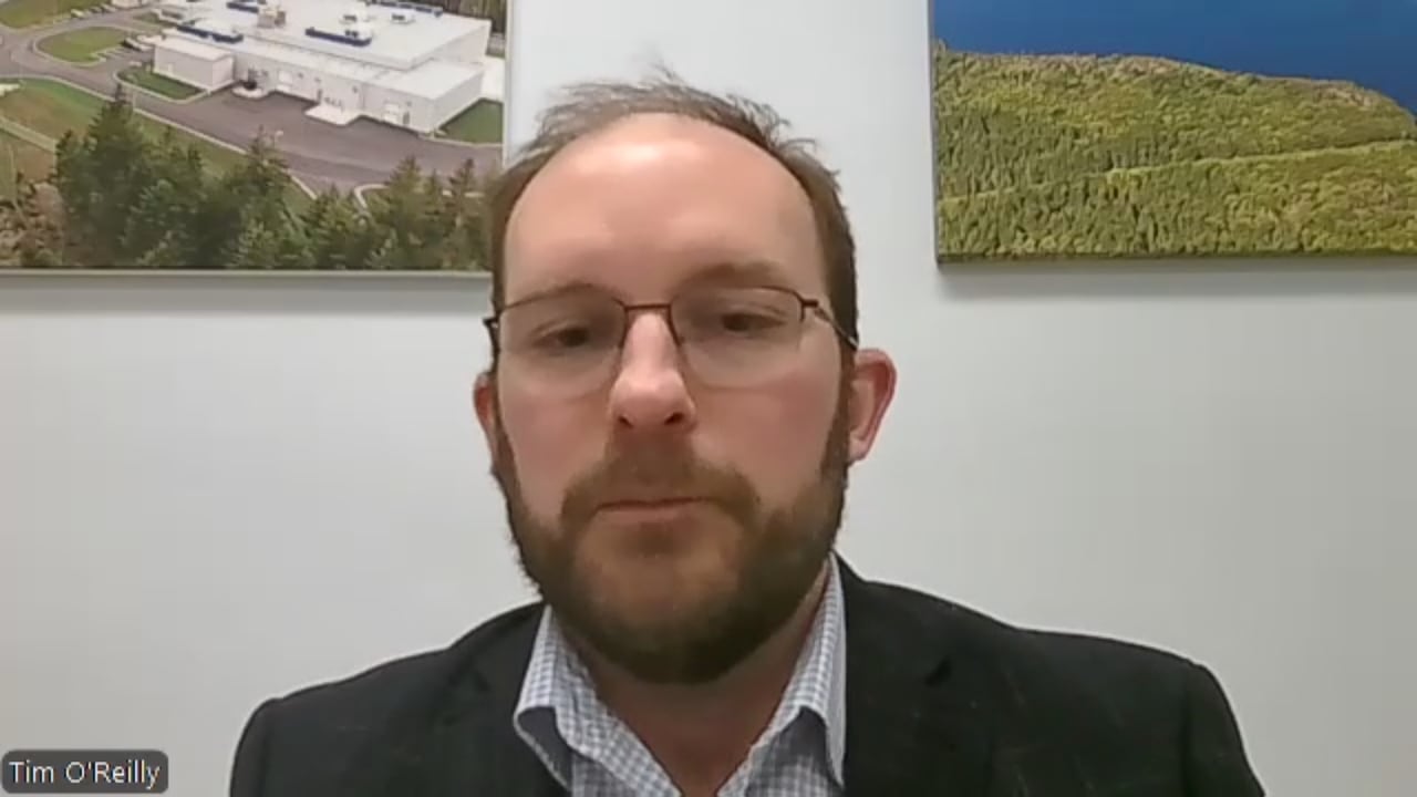 A man in a black suit jacket with a checkered collared shirt underneath, he has reddish-brown hair and a beard.