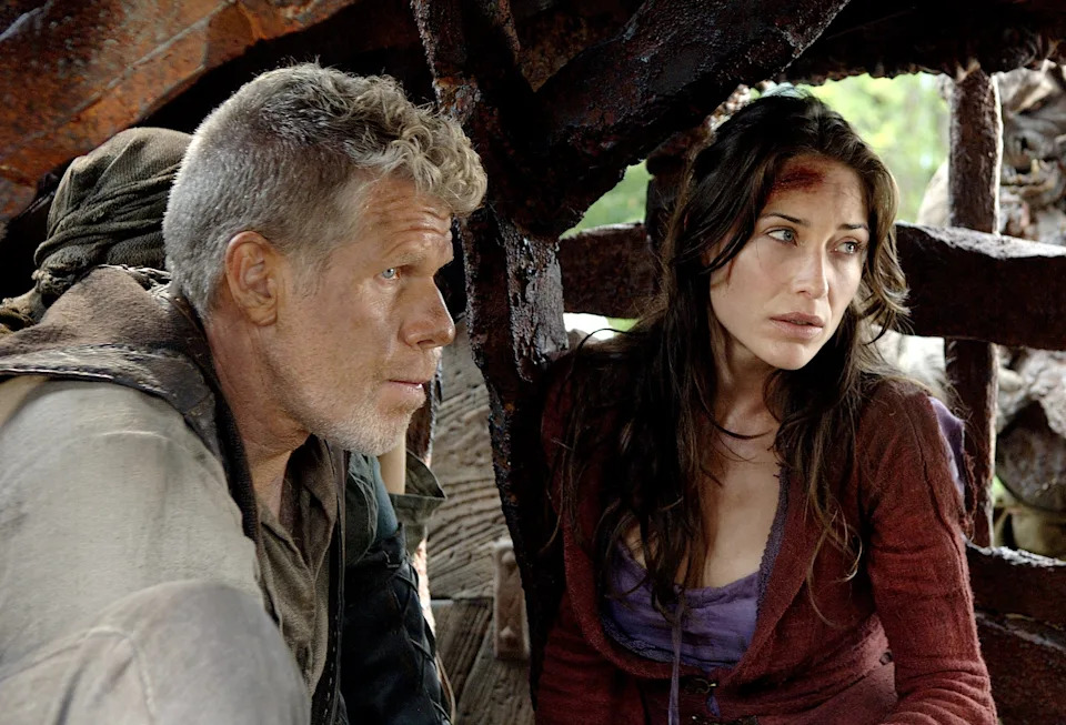 Two individuals, in survival-style clothing, are huddled under a weathered structure, appearing concerned, with a large creature nearby