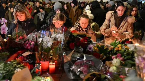 Getty Images People gather at the scene of a deadly bar fire in Switzerland. They are stood in front of a huge pile of flowers and candles.