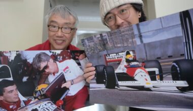 Two men smile at the camera while holding large photos of Formula 1 scenes, including a driver signing an autograph and a racing car on the track.
