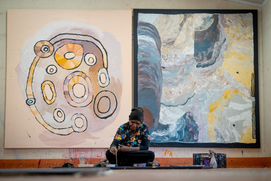 A woman sits on the floor painting with a stick. Two big paintings from the studio are on the wall behind her. 