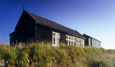 A wide shot of the radio station. It is a brown single-storey hut surrounded by grass.