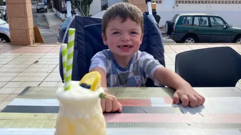 Tammy McDaid A young boy with short brown hair is smiling while sitting at an outdoor table in a holiday resort. A yellow coloured cocktail with a slice of lemon and green and white striped paper straws is pictured at the other side of the table.